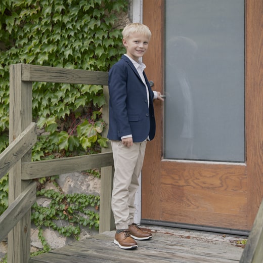 The image features a kid wearing a blue blazer, khaki pants, and brown Kids shoes while standing on a front stoop. Shop the Kids Shoes category now. 